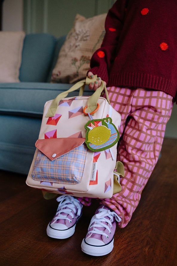 A child in patterned pink pants and lavender sneakers stands indoors, holding the Sticky Lemon Backpack Small - Special Edition - Snail Mail with geometric designs, a lemon-shaped keychain, and a waterproof lining. Only the lower body is visible.
