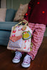A child in patterned pink pants and lavender sneakers stands indoors, holding the Sticky Lemon Backpack Small - Special Edition - Snail Mail with geometric designs, a lemon-shaped keychain, and a waterproof lining. Only the lower body is visible.