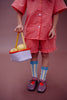 A child wearing a red checkered outfit holds a Sticky Lemon Treasure Bag - Dewy Lilac filled with lemons. They have blue and yellow striped socks, purple shoes, and stand against a brown background.