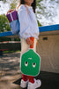 A young person stands near a concrete table outdoors, holding a paddle with a green face on it, part of their Sticky Lemon Pick Ball Game Set. They wear a light sweatshirt and white pants, with a colorful backpack over one shoulder and bright red socks with white sneakers. The background has trees and a fence.