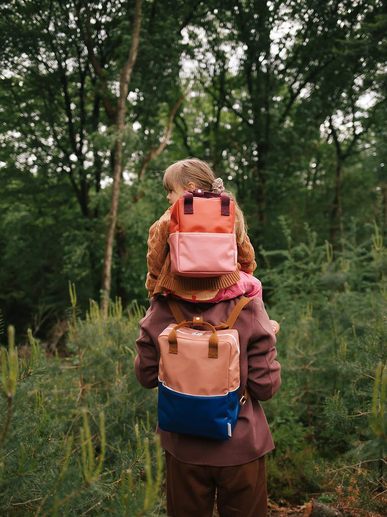 A young child with blonde braided hair, wearing a brown suit made from recycled PET bottles, carries a small child with a Sticky Lemon Backpack Large in Meet Me In The Meadows | Morning Sky on their shoulders amidst a lush green forest.