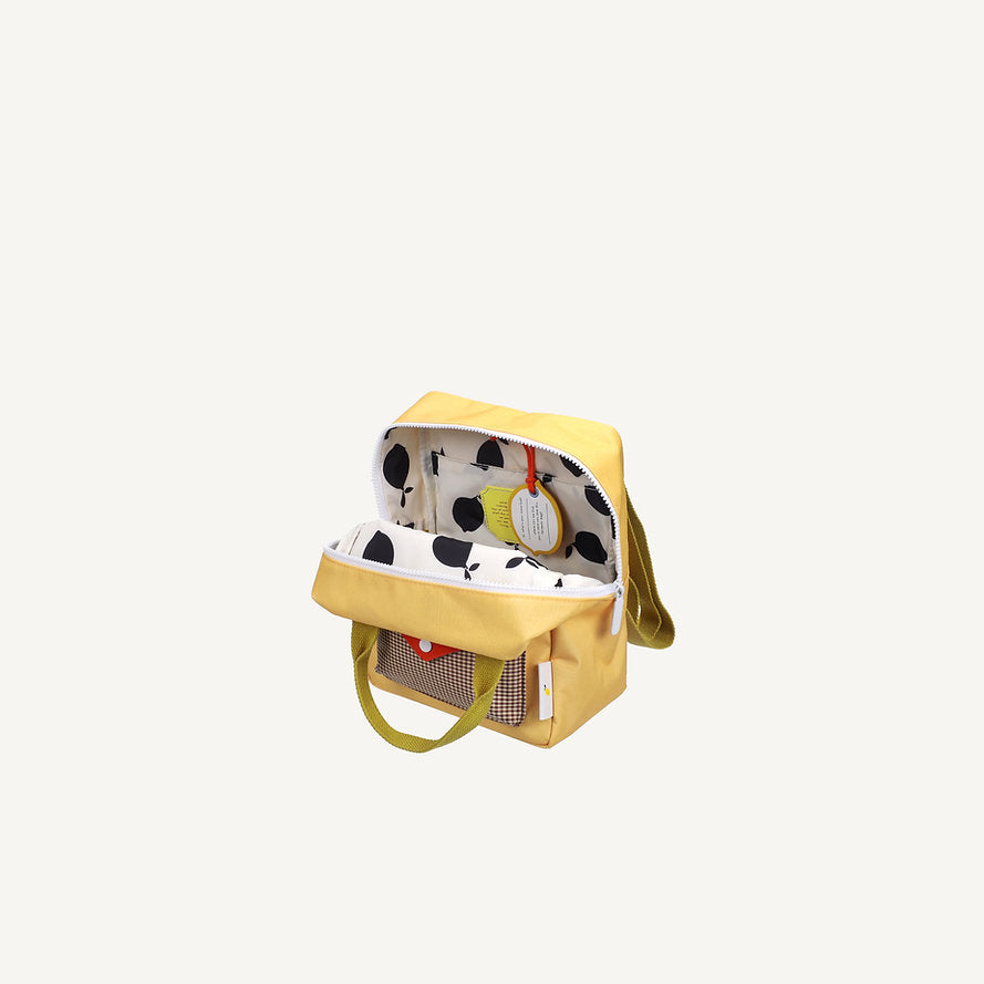 The Sticky Lemon Backpack Small - Envelope - Apple Juice is a yellow, rectangular kids backpack with green handles and a shoulder strap. It features a white interior with black fruit pattern, mesh pocket, and is made from waterproof recycled PET bottles.