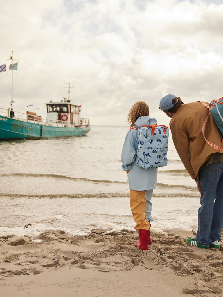 Two people stand on a sandy beach facing the water, a blue boat in the distance. One has a Sticky Lemon Backpack Large - Special Edition - Wonders of the Deep Sea, while the other wears a brown jacket and blue cap.