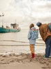 Two people stand on a sandy beach facing the water, a blue boat in the distance. One has a Sticky Lemon Backpack Large - Special Edition - Wonders of the Deep Sea, while the other wears a brown jacket and blue cap.