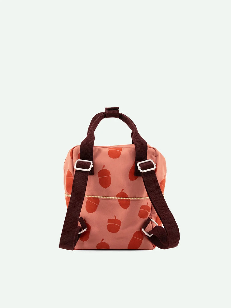 A small Sticky Lemon backpack with a peach background and a pattern of red apples. It features dark brown straps and silver buckles, with a zippered front pocket.