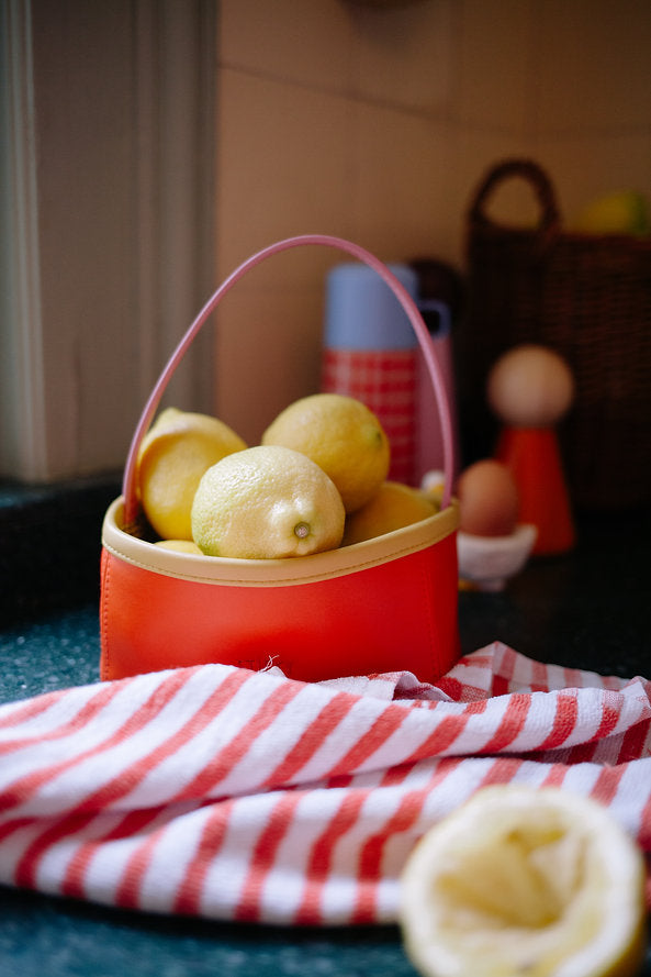 A red and yellow Sticky Lemon basket filled with lemons sits on a green countertop. In the background, kitchen items and a Sticky Lemon Treasure Bag in Campfire Orange are visible, with a red and white striped cloth in the foreground.