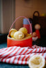 A red and yellow Sticky Lemon basket filled with lemons sits on a green countertop. In the background, kitchen items and a Sticky Lemon Treasure Bag in Campfire Orange are visible, with a red and white striped cloth in the foreground.