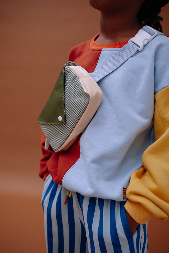 A person wearing a color-block sweatshirt, blue and white striped pants, and the Sticky Lemon Fanny Pack Small - Envelope - Tickling Grass stands against a plain brown background.
