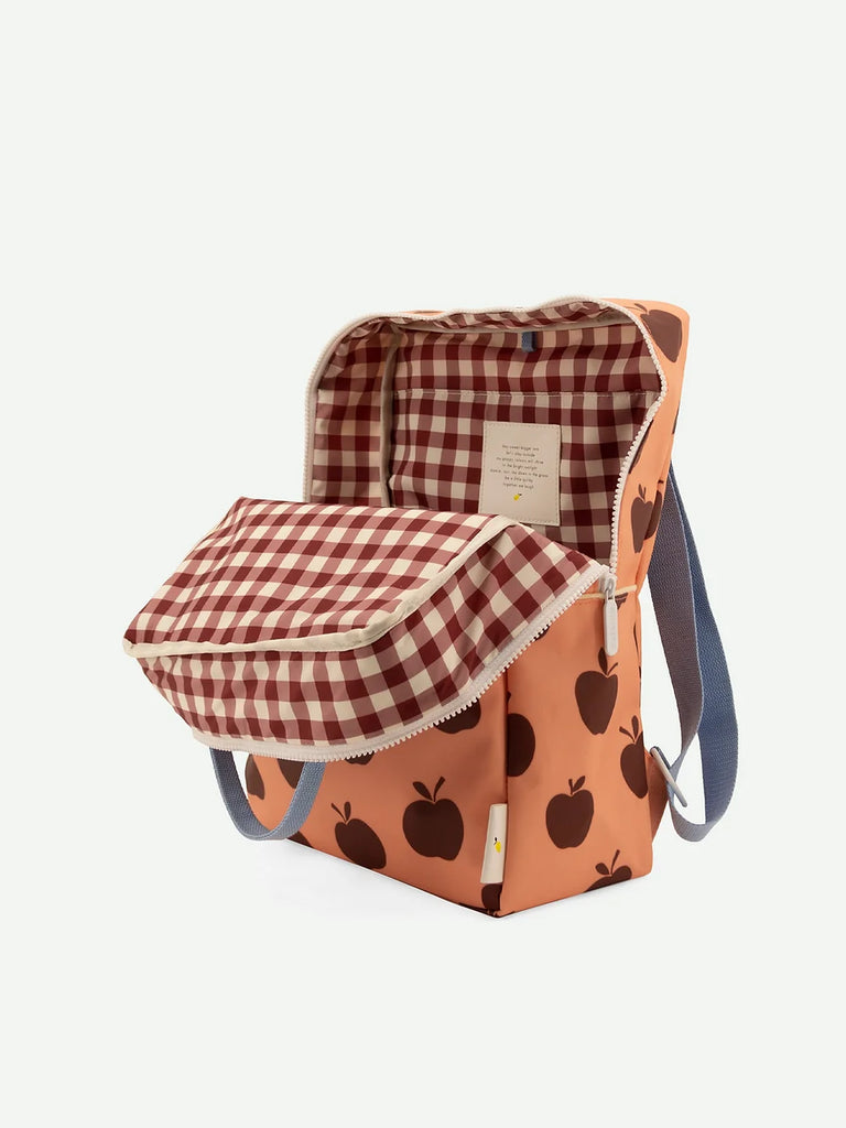 A peach-colored Sticky Lemon Backpack Large | Special Edition with a black apple pattern, featuring a blue strap and an open flap showing a red and white gingham interior with waterproof lining. The bag is positioned against a white background.