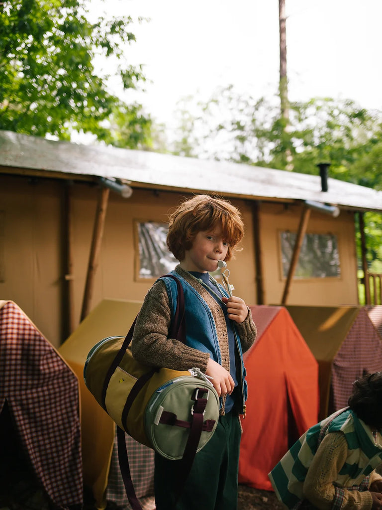 A young boy with curly red hair stands outside a tent, smiling and holding a marshmallow on a stick, ready for toasting. He carries a sleeping bag in a Sticky Lemon Duffle Bag | Adventure Collection | Khaki Green, dressed for.