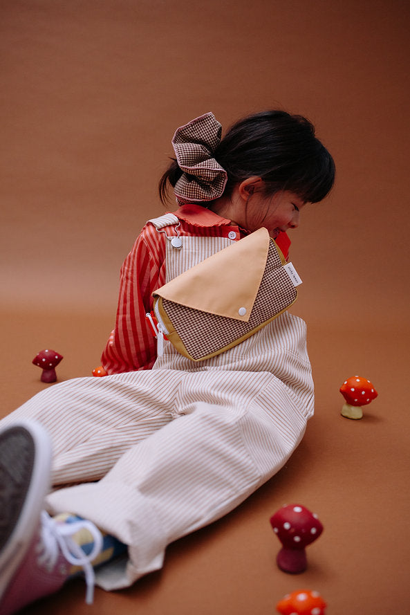 A young child in striped overalls and a red shirt sits on the floor among toy mushrooms, wearing a large checkered scrunchie and the Sticky Lemon Fanny Pack Small - Envelope - Apple Juice, against a brown background.