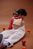 A young child in striped overalls and a red shirt sits on the floor among toy mushrooms, wearing a large checkered scrunchie and the Sticky Lemon Fanny Pack Small - Envelope - Apple Juice, against a brown background.