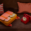 The Sticky Lemon School Bag in coral pink and submarine yellow, made from recycled PET with striped pockets, rests on a brown couch beside a red rotary phone; behind them are two red-and-white striped pillows.