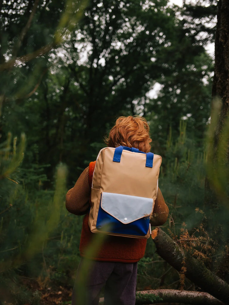 A young child with curly hair wearing a Sticky Lemon Backpack Large in Camp Yellow stands in a forest, viewed from behind, surrounded by lush green trees and underbrush.