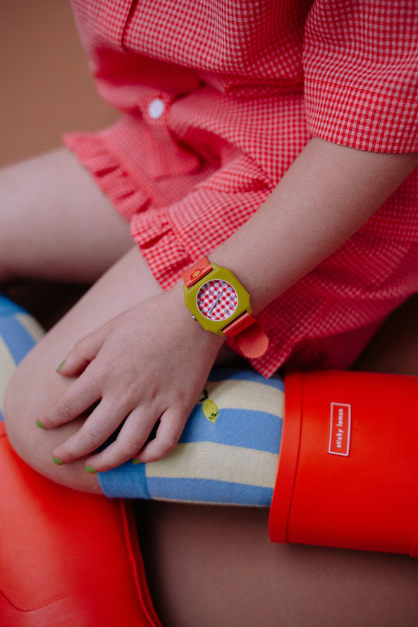 A child wears a red gingham dress, colorful striped socks, red rain boots, and the Sticky Lemon x Mini Kyomo Watch - Flower, which features a checkered face. Their nails are painted green.