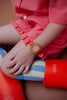 A child wears a red gingham dress, colorful striped socks, red rain boots, and the Sticky Lemon x Mini Kyomo Watch - Flower, which features a checkered face. Their nails are painted green.