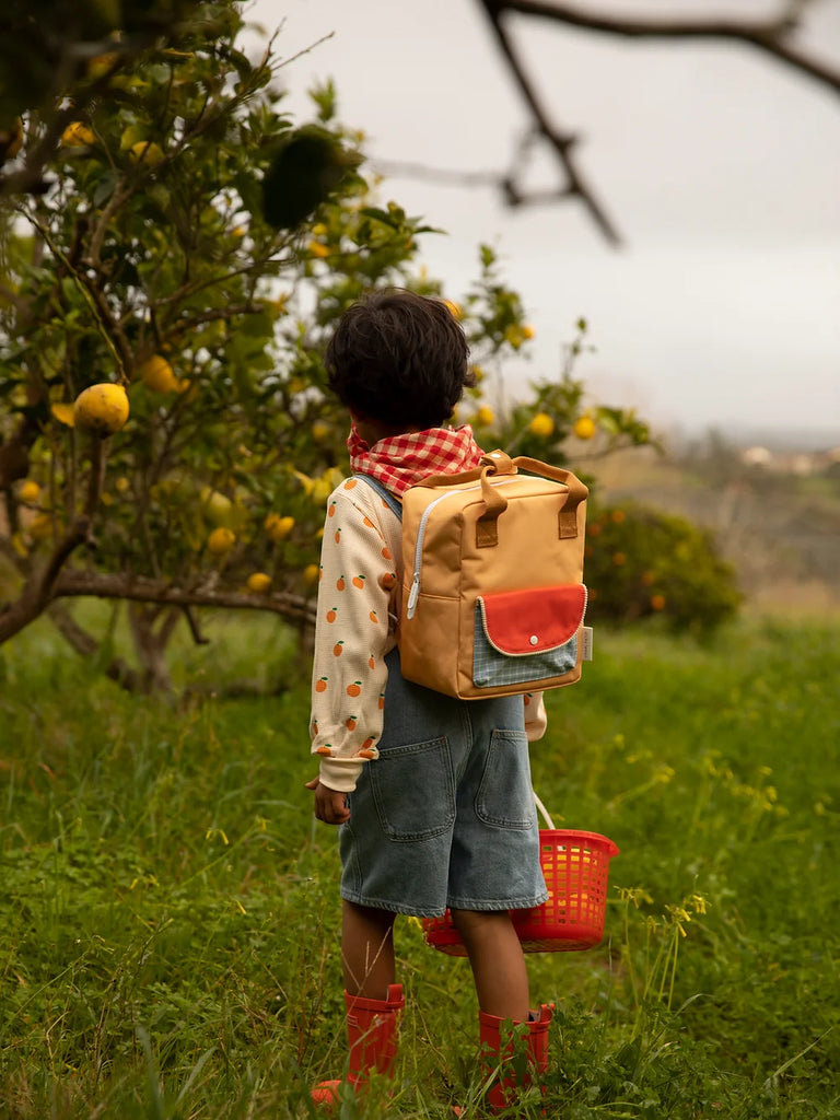 A young child wearing red boots and a Sticky Lemon Backpack Small made from recycled PET bottles stands in a lush orchard, facing away, looking up at the fruit-laden lemon trees, with a red bucket by.