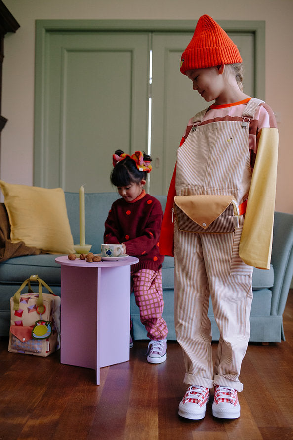 Two kids in a cozy living room wear colorful outfits. One wears overalls, a red beanie, and the Sticky Lemon Fanny Pack Small - Envelope - Apple Juice (made from recycled PET bottles); the other sits on a couch in plaid pants and a bow.