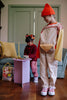 Two kids in a cozy living room wear colorful outfits. One wears overalls, a red beanie, and the Sticky Lemon Fanny Pack Small - Envelope - Apple Juice (made from recycled PET bottles); the other sits on a couch in plaid pants and a bow.