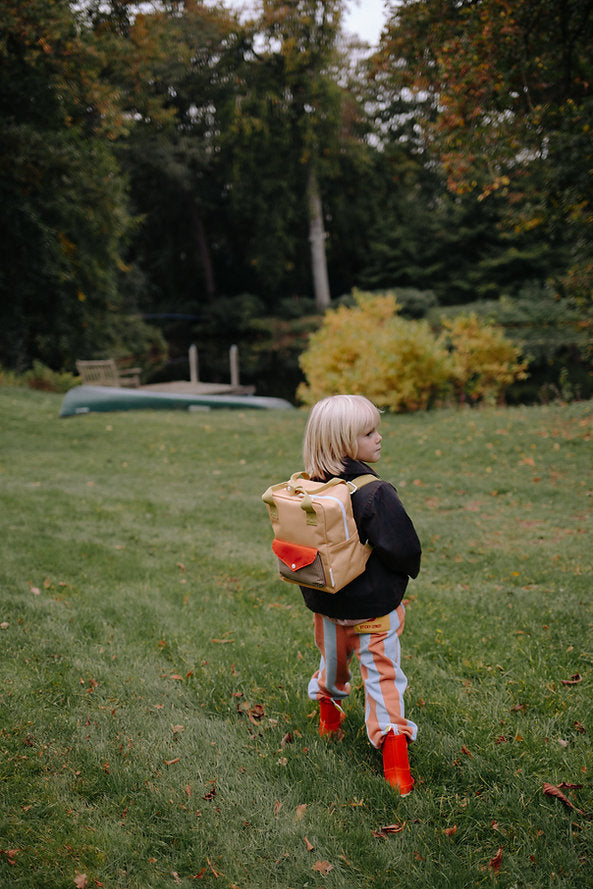 A young child with blonde hair, wearing a Sticky Lemon Backpack Small - Envelope - Apple Juice, striped pants, and red boots, stands on a grassy lawn with trees and a canoe in the background.