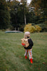 A young child with blonde hair, wearing a Sticky Lemon Backpack Small - Envelope - Apple Juice, striped pants, and red boots, stands on a grassy lawn with trees and a canoe in the background.