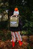 A child in a colorful beanie, striped pants, and red boots stands in a leaf-covered forest with their back to the camera, carrying the Sticky Lemon Backpack Small - Envelope - Tickling Grass made from recycled PET bottles.