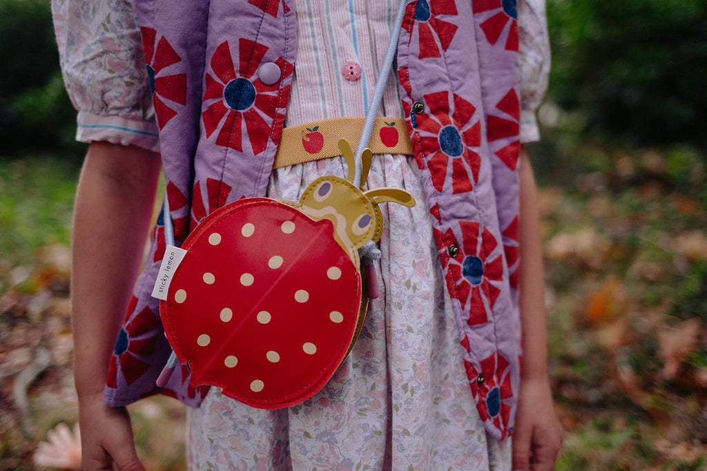 A child outdoors wears a purple vest with red patterns, a floral dress, and a yellow belt with red apples, carrying the playful Sticky Lemon Wallet Bag - Ladybug.