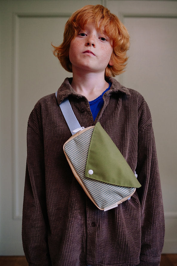 A young red-haired person in a brown corduroy shirt wears the Sticky Lemon Fanny Pack Small - Envelope - Tickling Grass, made from recycled PET bottles, standing before a light wall and looking at the camera.