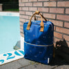 The Sticky Lemon Backpack Large | Special Edition Eyes | Boxing Blue—featuring tan handles, a front pocket, and a cute white smiley face pattern made from recycled PET bottles—rests on the ground near the edge of a swimming pool with a brick wall in the background.