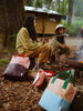 Two people roasting marshmallows over a campfire in a forest, with Sticky Lemon Backpack Large | Color Blocking | Meet Me In The Meadows | Moonrise Pink backpacks crafted from recycled PET bottles nearby, depicting a serene outdoor camping scene.