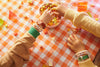 Two people with beaded bracelets reach for colorful snacks on an orange checkered tablecloth, both wearing a Mini Kyomo Watch - Cinnamon Roll. Scattered snacks and geometric shapes decorate the table.