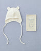 A white Baby Pilot Hat made from organic cotton with bear ears and long strings sits next to a drawstring pouch on a light blue background. The pouch has an illustration of the hat and the text "baby pilot hat 0-3 months" printed on it.