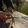 A person whittles a stick with the Huckleberry Pocket Knife (Not a Toy) on a wooden bench outdoors, sunlight shining and a blurred grassy field in the background.