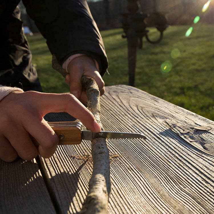 A person whittles a stick with the Huckleberry Pocket Knife (Not a Toy) on a wooden bench outdoors, sunlight shining and a blurred grassy field in the background.