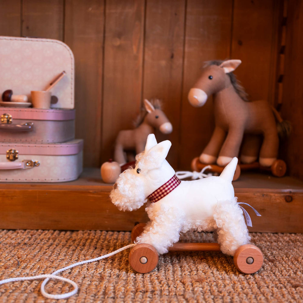 Pull-Along Lola, a white plush dog toy on wheels with a string, sits on a woven rug. Two stacked toy suitcases and two plush horse toys are behind it—a perfect toddler gift for imaginative developmental play.