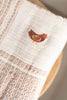 A close-up of the Organic Cotton Baby Blanket - Farm shows its beige, textured fabric with a brown embroidered chicken, resting on a wooden surface.