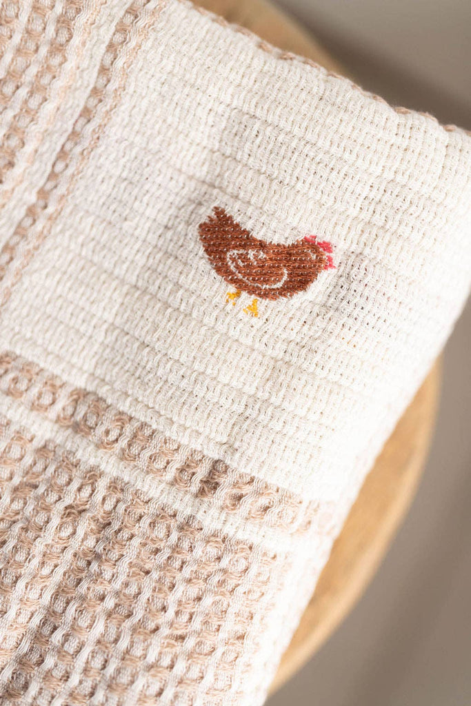A close-up of the Organic Cotton Baby Blanket - Farm shows its beige, textured fabric with a brown embroidered chicken, resting on a wooden surface.