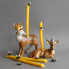 Two ceramic deer figurines with party hats display yellow candles from the Beeswax Birthday Candle Set on their backs. Extra beeswax candles and tiny bee decorations are scattered nearby, all set against a plain gray background.