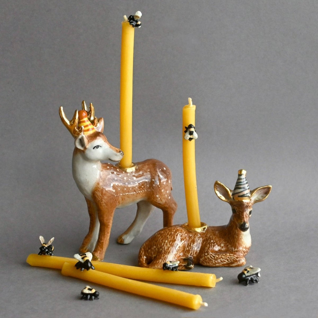 Two ceramic deer figurines with party hats display yellow candles from the Beeswax Birthday Candle Set on their backs. Extra beeswax candles and tiny bee decorations are scattered nearby, all set against a plain gray background.