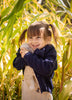 A young girl with brown pigtails, wearing a navy sweater, smiles as she hugs her Senger Naturwelt Cuddly Animal - Linen Sheep, standing among tall green plants in warm sunlight.