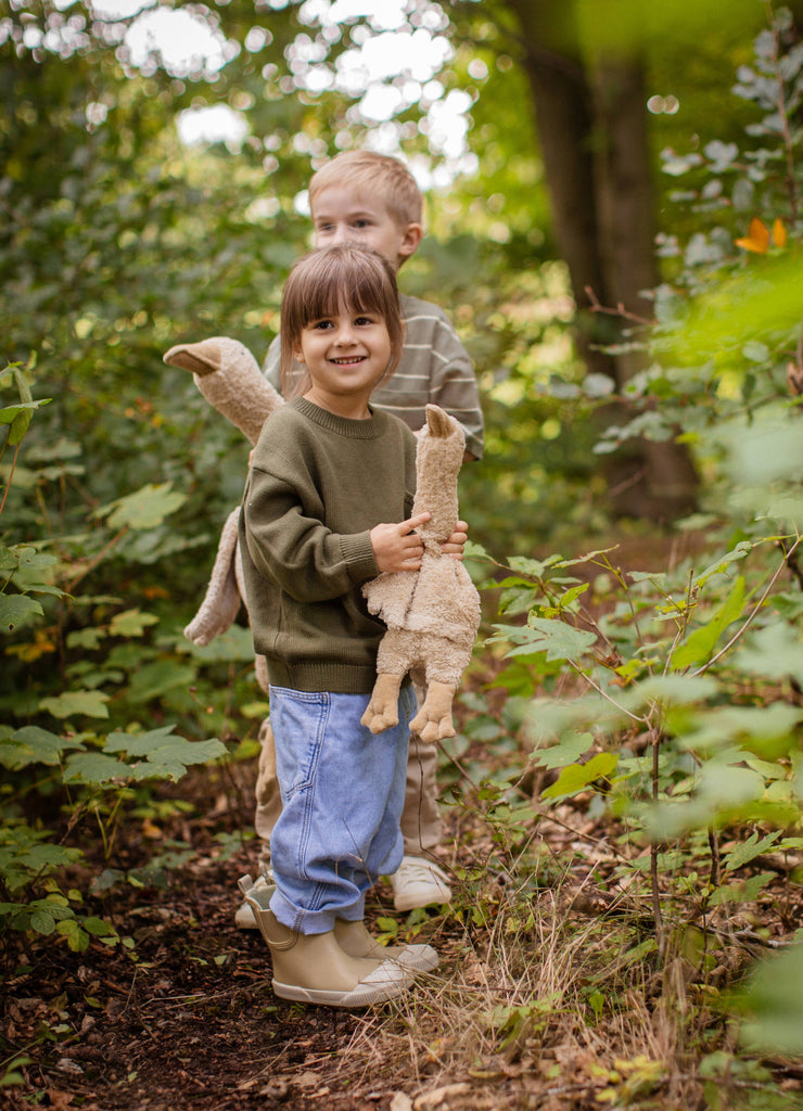 Two young children stand on a forest path surrounded by greenery. The front child smiles while holding a Senger Naturwelt Cuddly Animal - Linen Goose; the child behind wears a striped shirt. Both look happy and enjoy nature.