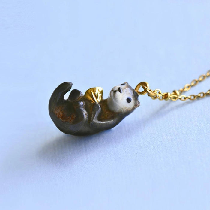 A small, whimsical River Otter "Golden Gift" Necklace, hand painted and hanging from a gold chain, displayed against a pale blue background. The otter is depicted in mid-swim with a playful