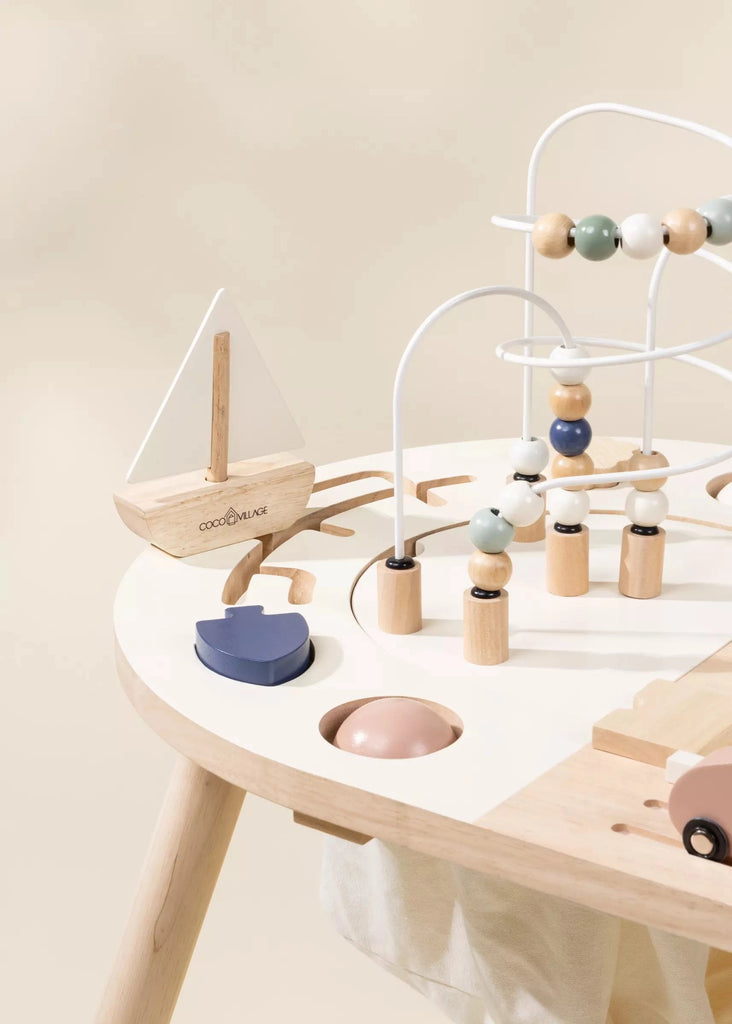 A modern, minimalist Wooden Activity Table featuring various geometric toys, including a bead maze and a sailing boat, on a soft beige background.