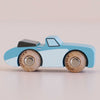 A small blue wooden toy car, the No. 1 GT10 Justin made in England, featuring natural wood wheels and a black rear spoiler reminiscent of a classic car, is placed on a light surface.