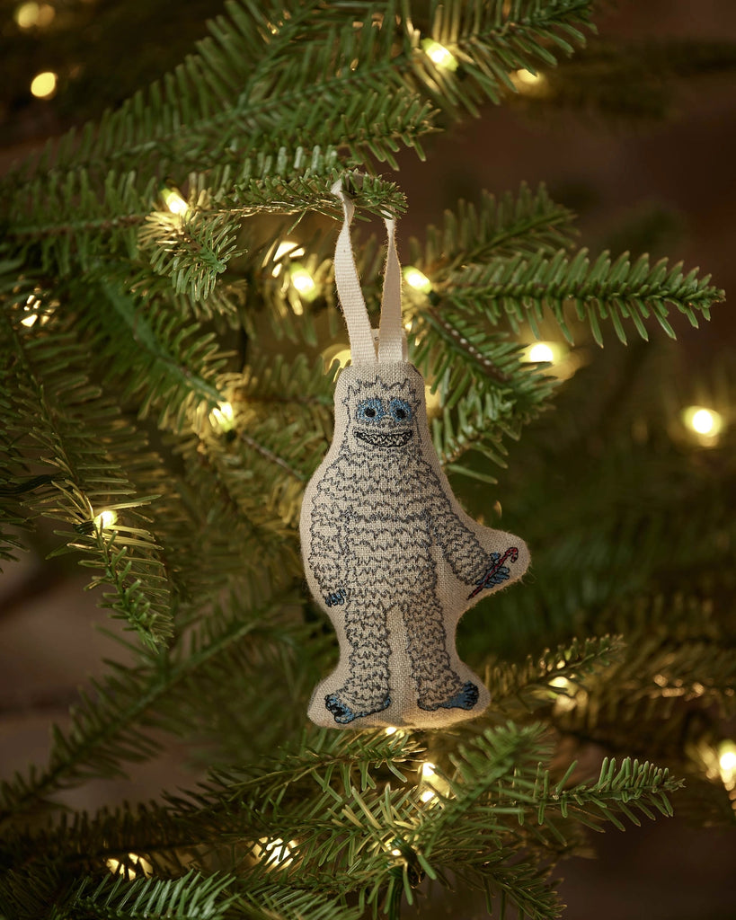 The Coral & Tusk Yeti with Candy Cane Ornament, featuring embroidered details, hangs on a green Christmas tree branch with glowing white lights—a charming holiday decoration and perfect Christmas gift.