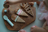 A child uses the Ice Cream Cutter Set - Eco Playdough Tool to decorate ice cream cone-shaped creations with pink and white playdough and colorful sprinkles on a wooden board, surrounded by cookie cutters and eco-friendly tools, creating a delightful sensory play experience.