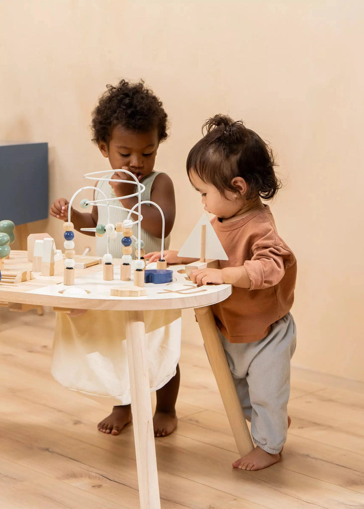 Two toddlers, one with curly hair and another with straight hair, are playing with a Wooden Activity Table at a small activity table in a warmly lit room.