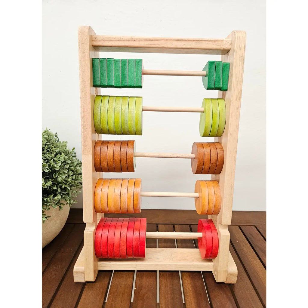 The Fruitful Abacus, a wooden educational tool designed to enhance counting skills, is shown with five rows of vibrant discs. These discs are arranged in descending order of green, light green, yellow, orange, and red. The abacus stands on a wooden surface with a partial view of a potted plant on the left side.