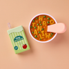 The Tiny Tummies Set - Alphabet Soup Bowl and Juice Box For Dolls features a bowl of alphabet soup with peas and a pink spoon, alongside a green juice box labeled “tiny tummies apple juice,” all set against a peach background. Perfect for play sets or as doll accessories, the soup contains letter shapes including H, R, O, T, A, N, I, and W.