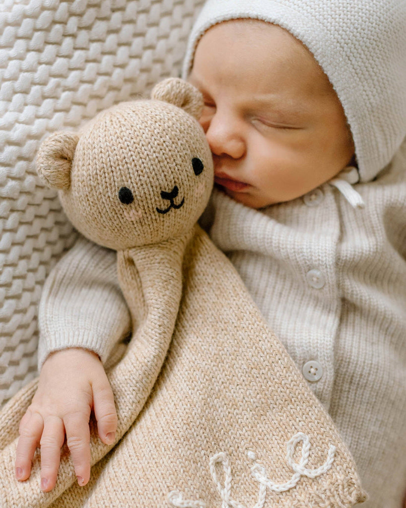 A baby sleeps with the Cuddle + Kind Organic Cotton Baby Bear Lovey (Honey), cuddled under a soft blanket.
